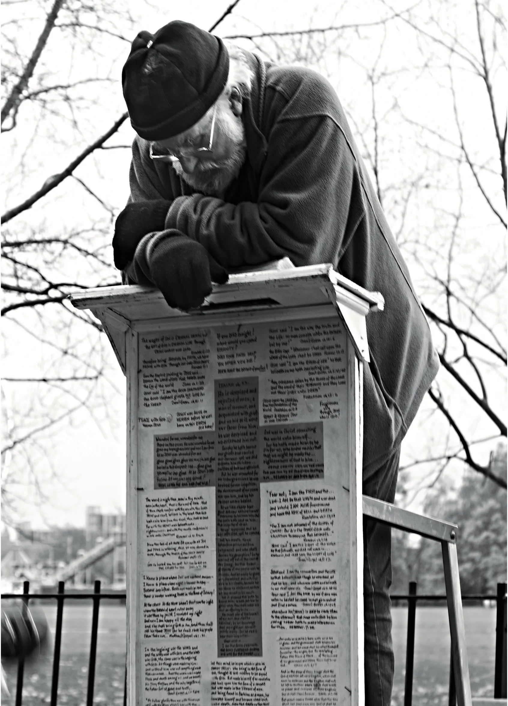 A speaker in a cap leaning over a small lectern covered in hand-written notes, bare trees behind him.