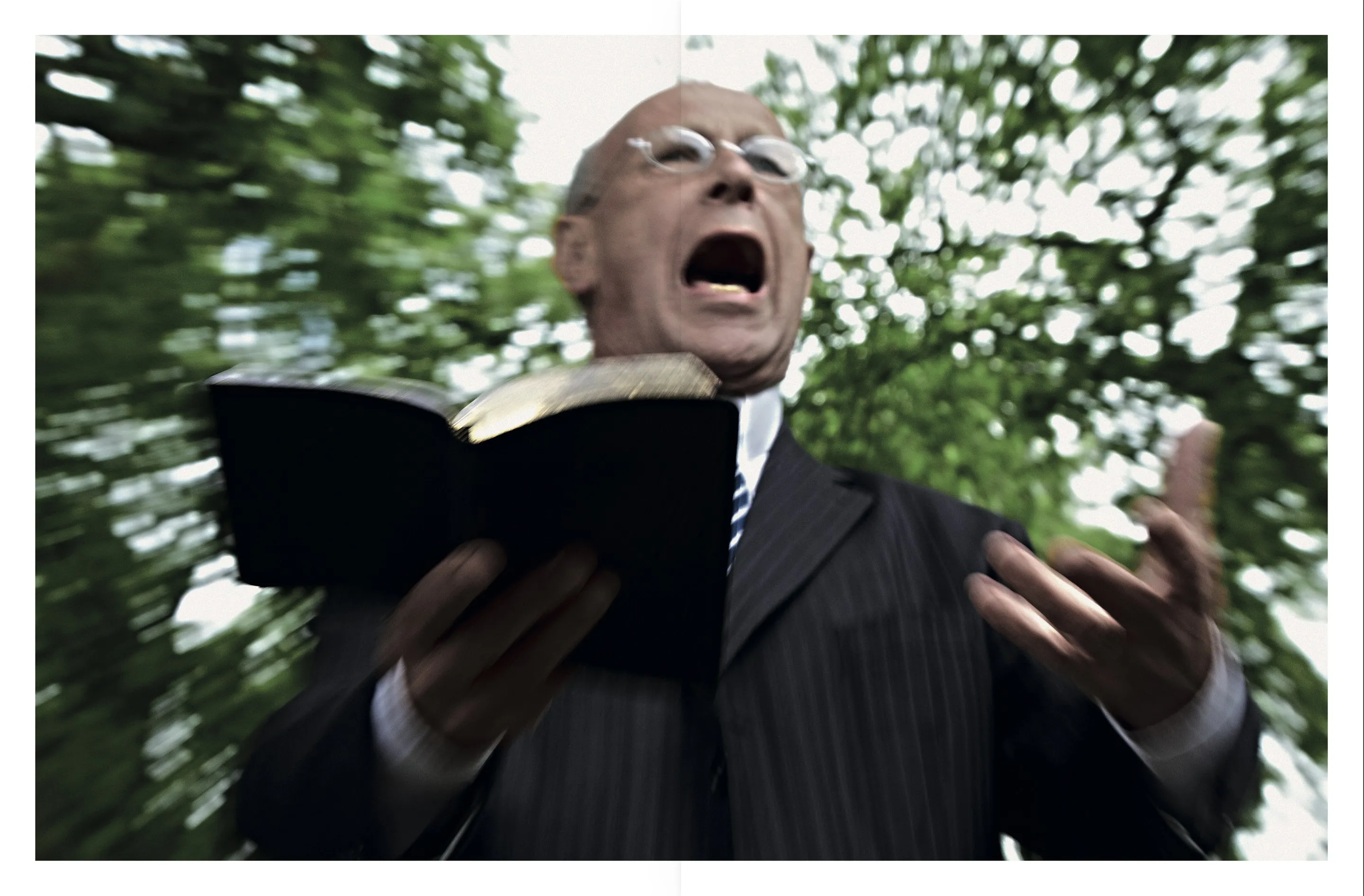 A preacher in a pinstripe suit holding an open book and raising his hand, with blurred green trees behind him.