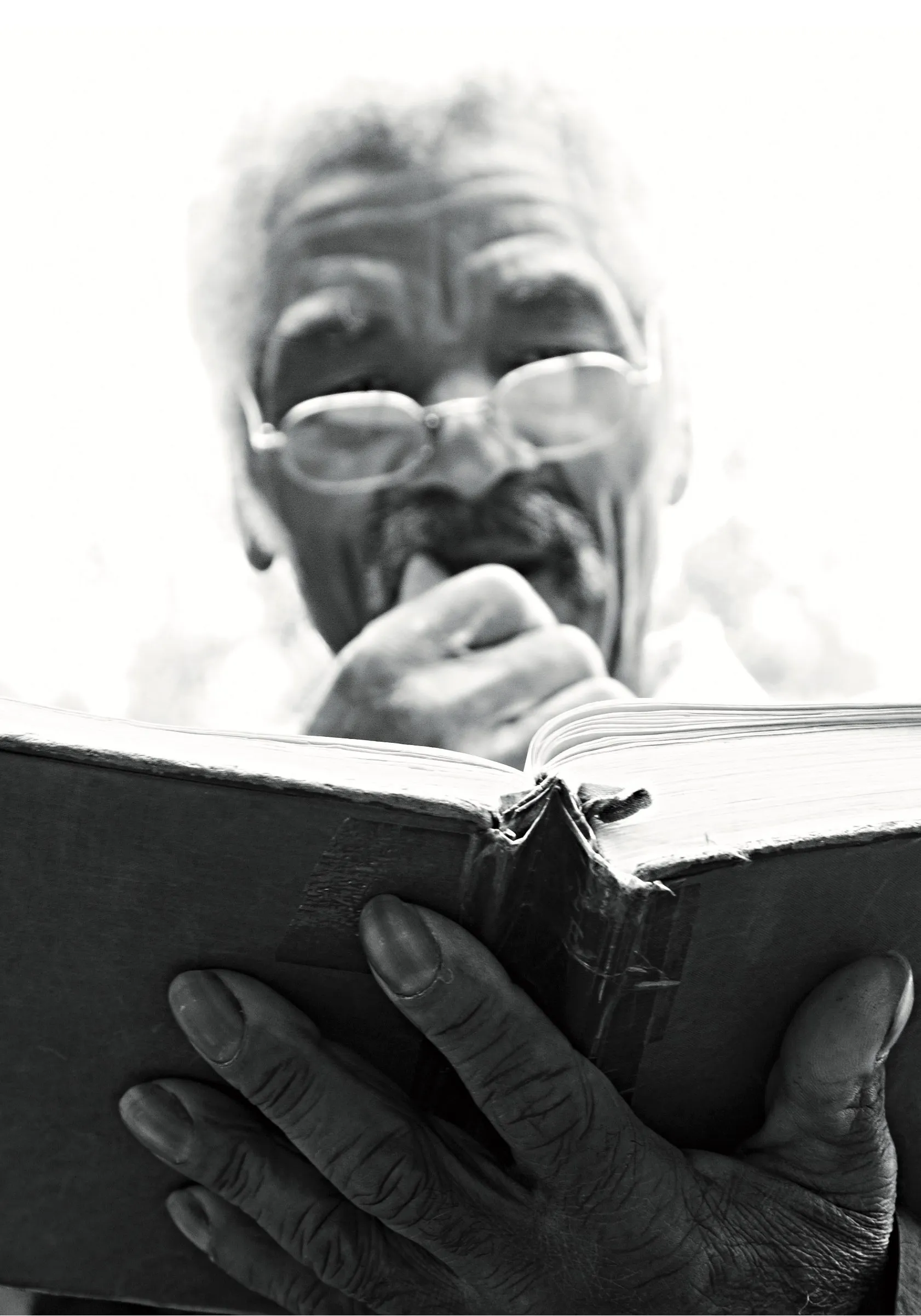 Black and white close-up of an elderly man with glasses reading from an open book, seen from below.