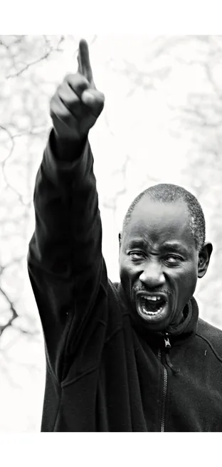 Black and white photograph of a man shouting and pointing his finger to the sky, bare tree branches behind him.